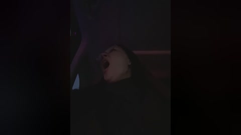 Media: A dimly lit video captures a close-up of a young woman with long black hair, lying on her back, mouth open in apparent distress. The background is dark and blurred, focusing on her face.