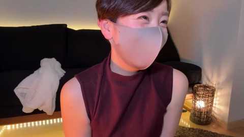 Media: Video of an Asian woman with short brown hair, wearing a sleeveless maroon top and a face mask, seated indoors, near a black couch with a white blanket, a lit candle, and a decorative lantern on a wooden floor.