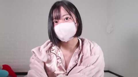 Media: Video of an Asian woman with short black hair, wearing a white surgical mask, light pink satin robe, and sitting in a plain white room with a blue and red object in the background.