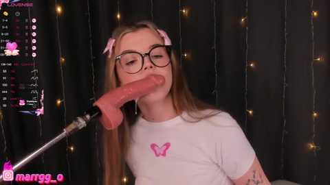 Media: A video shows a young woman with glasses, wearing a pink bow headband and a white t-shirt with a butterfly, sucking a large, realistic, pink dildo. Background features dark curtains with string lights.