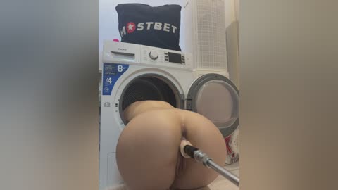 Media: A video of a nude woman, with her buttocks prominently displayed, cleaning a white washing machine with a hose in a laundry room.