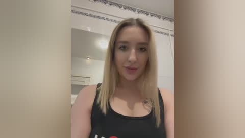 Media: Video of a young woman with fair skin, long blonde hair, and a slender build. She wears a black tank top. Background features a white bathroom with decorative molding and a mirror.