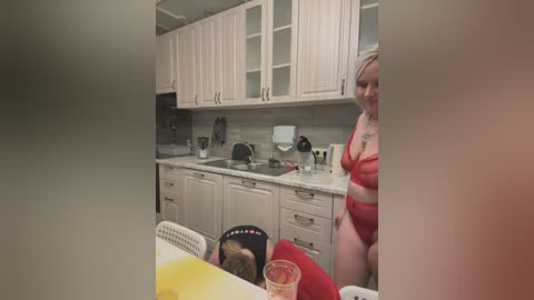 Media: A video captures a woman in red lingerie standing in a modern kitchen, smiling, while a man in a red shirt sleeps at a table.