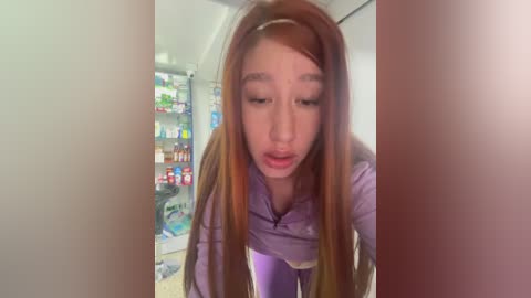 Media: Video of a young Asian woman with long, straight, auburn hair, wearing a purple hoodie, looking down into an open refrigerator filled with various food items.