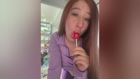 Media: Video of a young, fair-skinned woman with long, straight red hair, wearing a purple top, licking a red lollipop. The background shows a modern kitchen with a fridge filled with colorful food items.