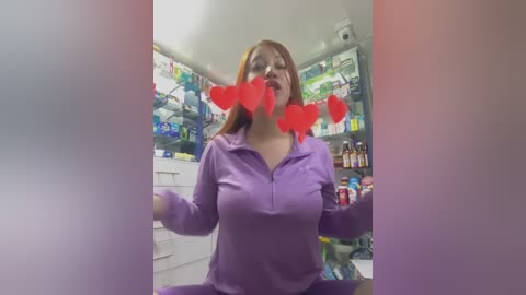 A video of a woman with red hair, wearing a purple hoodie, standing in a brightly lit, organized pantry filled with colorful products, with red heart emojis overlaying her face.