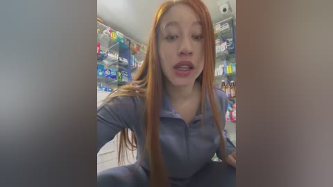 A video of a young, pale-skinned woman with long, straight, auburn hair, wearing a blue jumpsuit, sitting in a brightly lit pharmacy. She has a neutral expression and is surrounded by colorful shelves of medication.