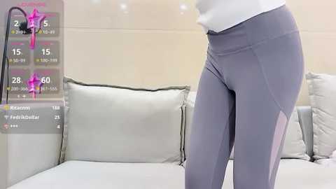 Video of a person wearing form-fitting, light purple leggings and a white top, standing in a modern, minimalistic living room with beige walls and a white sectional sofa.