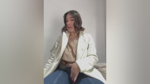 Media: A video of a young woman with medium-length, wavy dark hair, wearing a beige sweater, blue jeans, and a white coat, sitting on a beige couch against a plain white wall.