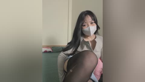 Media: Video of a young Asian woman with long black hair, wearing a gray face mask, gray blouse, and black stockings, crouching behind a pillar, revealing a hint of her bare thigh.