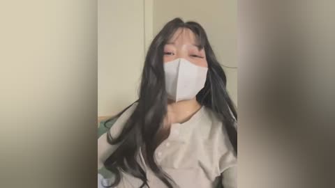 Media: A video of an Asian woman with long black hair and fair skin, wearing a white surgical mask, beige top, and light green wall background.