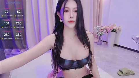 Media: Video of an East Asian woman with long black hair, wearing a black leather bra, sitting in a room with a white bed, flowers, and a video game screen showing stats.