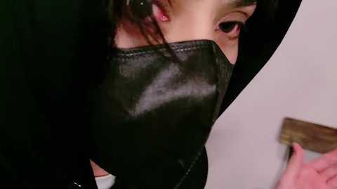 Media: Video of a person wearing a black leather mask with pink lipstick, eyes partially visible, holding a wooden door handle, suggesting an indoor setting.