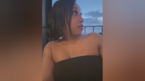 Media: Video of a dark-skinned woman with short black hair, wearing a black strapless dress, gazing out a window at a sunset sky.