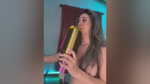 Media: Video of a topless woman with long, wavy hair, drinking from a colorful, multicolored water bottle. Background features a maroon curtain, a TV, and a greenish-blue light.