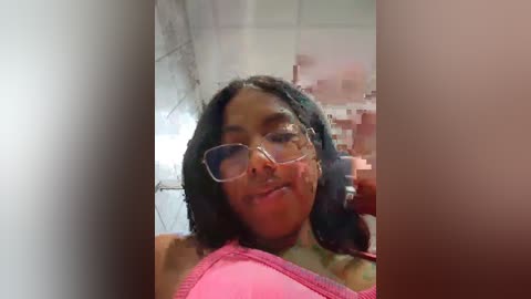 Media: A video of a young Black woman with medium brown skin, wearing glasses and a pink bra, smiling, taken in a bathroom with a white tiled wall and a mirror.