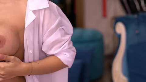 Media: A close-up video of a woman with light skin and medium-sized breasts, partially covered by an unbuttoned white shirt, holding her breast. Background includes a teal chair and blurred objects.