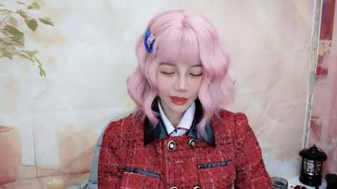 Media: Video of a young woman with pastel pink hair and a blue hair clip, wearing a textured red blazer and a white shirt, sitting in front of a soft-focus, floral-patterned background.