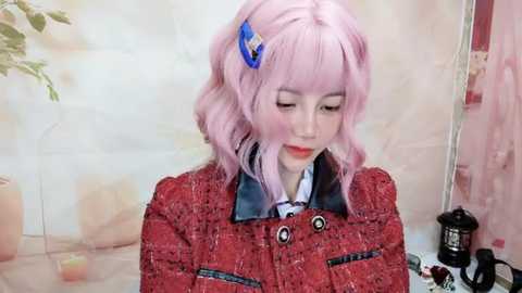 Media: Video of a young woman with pale skin and pink, wavy hair styled with a blue hair clip. She wears a red textured blazer over a blue shirt, with a potted plant and a black kettle in the background.