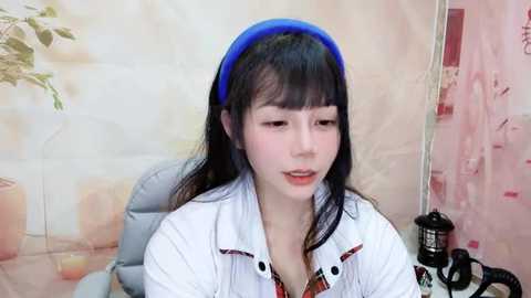 Media: Video of an East Asian woman with long black hair and fair skin, wearing a blue headband and white shirt, sitting on a grey chair, in a pastel-colored room with potted plants.