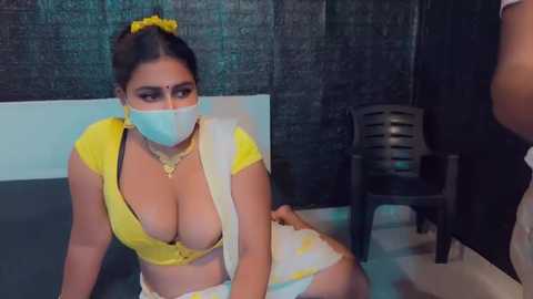 Media: Video of a young South Asian woman with medium skin tone, wearing a yellow crop top revealing ample cleavage, a white hijab, and a face mask. She sits on a chair in a dimly lit room with dark walls.