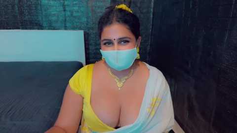 Media: Video of a South Asian woman with dark hair, wearing a yellow saree, white shawl, and blue face mask, seated on a bed with dark bedding, in a dimly lit room.