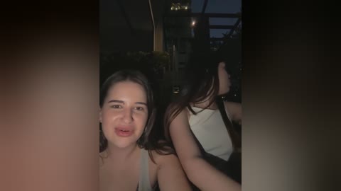 Media: Video of a young woman with fair skin, brown hair, and a light-colored tank top, smiling, and holding a phone, in a dimly lit, possibly outdoor restaurant setting.