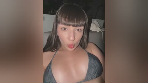 Media: Video of a young woman with straight dark hair and blunt bangs, wearing a dark blue bikini top, making a surprised face, indoors with blurred background.