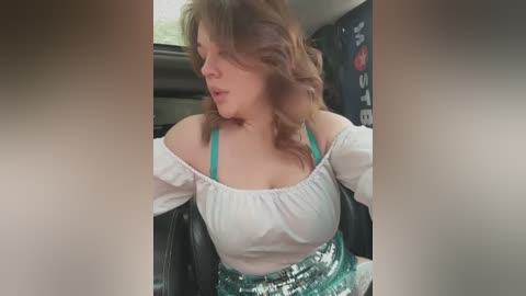 Media: Video of a Caucasian woman with wavy brown hair, wearing a white off-shoulder blouse and green sequined skirt, sitting in a car.