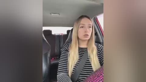 Media: Video of a young woman with long blonde hair, wearing a striped shirt, seated in a car with a seatbelt on. The car interior is dark with red and black accents.