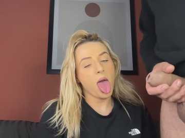 Media: Video of a blonde woman with fair skin, wearing a black t-shirt, sticking out her tongue and touching a man's erect penis. Background features a red wall and abstract art.