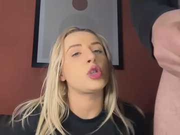 Media: Video of a blonde woman with light skin, wearing a black top, puckering her lips in an intimate kiss with a man's torso. Background features a framed abstract painting on a red wall.