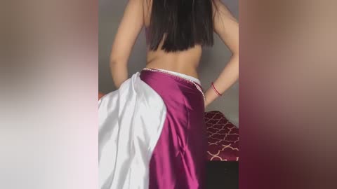 Media: A video of a woman with long, straight black hair, wearing a purple and white skirt, standing in a bedroom with a red patterned bedspread.