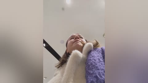 Media: A video capturing a close-up view of a woman with brown hair, wearing a white coat and a lavender cardigan, looking up with a serene expression, in a softly lit room.