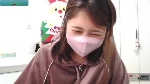 Video of an Asian woman with straight, shoulder-length dark hair, wearing a pink face mask, a light brown hoodie, and a white t-shirt. She has her eyes closed and appears to be coughing. Background shows a white wall with festive holiday decorations.