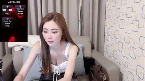 Media: Video of a young Asian woman with long brown hair in a white bra, sitting on a beige sofa with geometric-patterned wallpaper in the background.