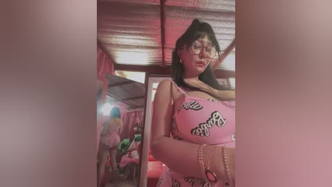 Media: Video of a woman with long black hair, wearing a pink crop top with \"love\" print, standing in a dimly lit, rustic room with a wooden ceiling and a mirror reflecting her.