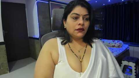 Media: Video of a middle-aged Indian woman with medium skin tone, wearing a white sleeveless top, sitting in a modern bedroom with a bed and dark curtains.