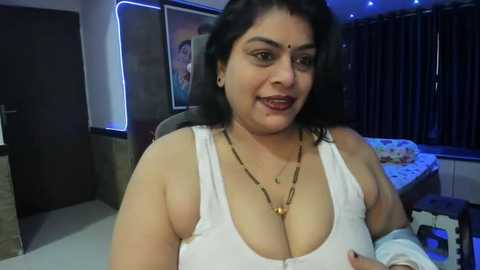 Media: Video of a South Asian woman with medium skin tone, dark hair, and a confident smile, wearing a revealing white tank top, sitting in a dimly lit bedroom with dark curtains and a bed in the background.