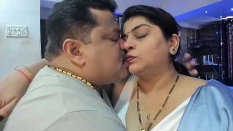 Media: Video of an Indian couple, both with light brown skin, in a cozy indoor setting. The man, with short dark hair, is kissing the woman, who has short dark hair and is wearing a blue sari.