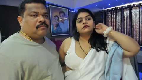 Media: Video of an overweight South Asian man with a mustache in a white shirt, next to a plus-size woman with dark hair, in a modern living room with framed portraits and patterned curtains.