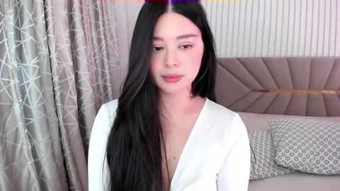 Media: Video of a young Asian woman with long, straight black hair, wearing a white robe, standing in a modern bedroom with a tufted headboard, grey curtains, and patterned pillows.