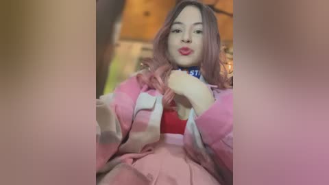 Video of an Asian woman with long, wavy pink hair and light skin, wearing a pink robe and red top, making a pouty face. Background is blurred with wooden textures.