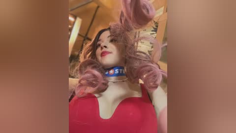 Media: Video of a woman with fair skin and long, curly, pink wig, wearing a red halter top and blue collar. She's indoors, with blurred background.