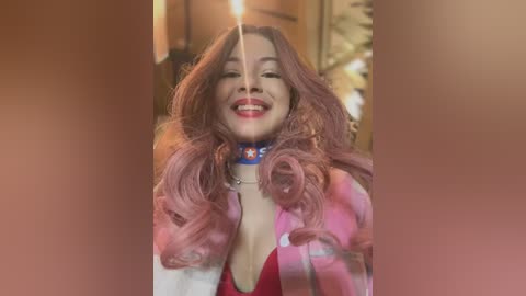 Video of a young woman with long, wavy pink hair, wearing a red top and blue collar, smiling widely with a lit candle in her mouth, indoors.