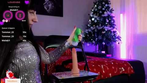 Media: Video of a woman with long black hair and green hair extensions, wearing a glittery silver dress, holding a large pink dildo, in a bedroom with a Christmas tree and red bedspread.