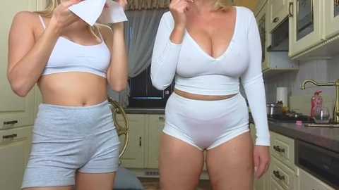 Media: Video of two blonde women in white sports bras and shorts, one holding a paper, in a modern kitchen.