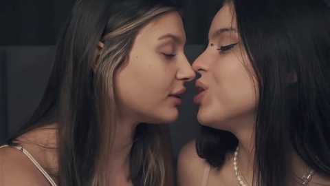 Media: Video of two young women with long dark hair, wearing light-colored tops, sharing a kiss on the lips, set against a dimly lit background.