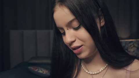 Media: A video of a young woman with long, dark hair, light skin, and a serene expression, wearing a pearl necklace. She is indoors with a blurred, dark background.