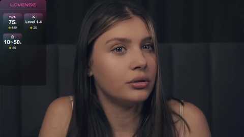 Media: Video of a young woman with light brown skin, straight dark hair, and brown eyes, wearing a pink top, in a dimly lit room with a digital game overlay showing \"LoveMe\" and stats.